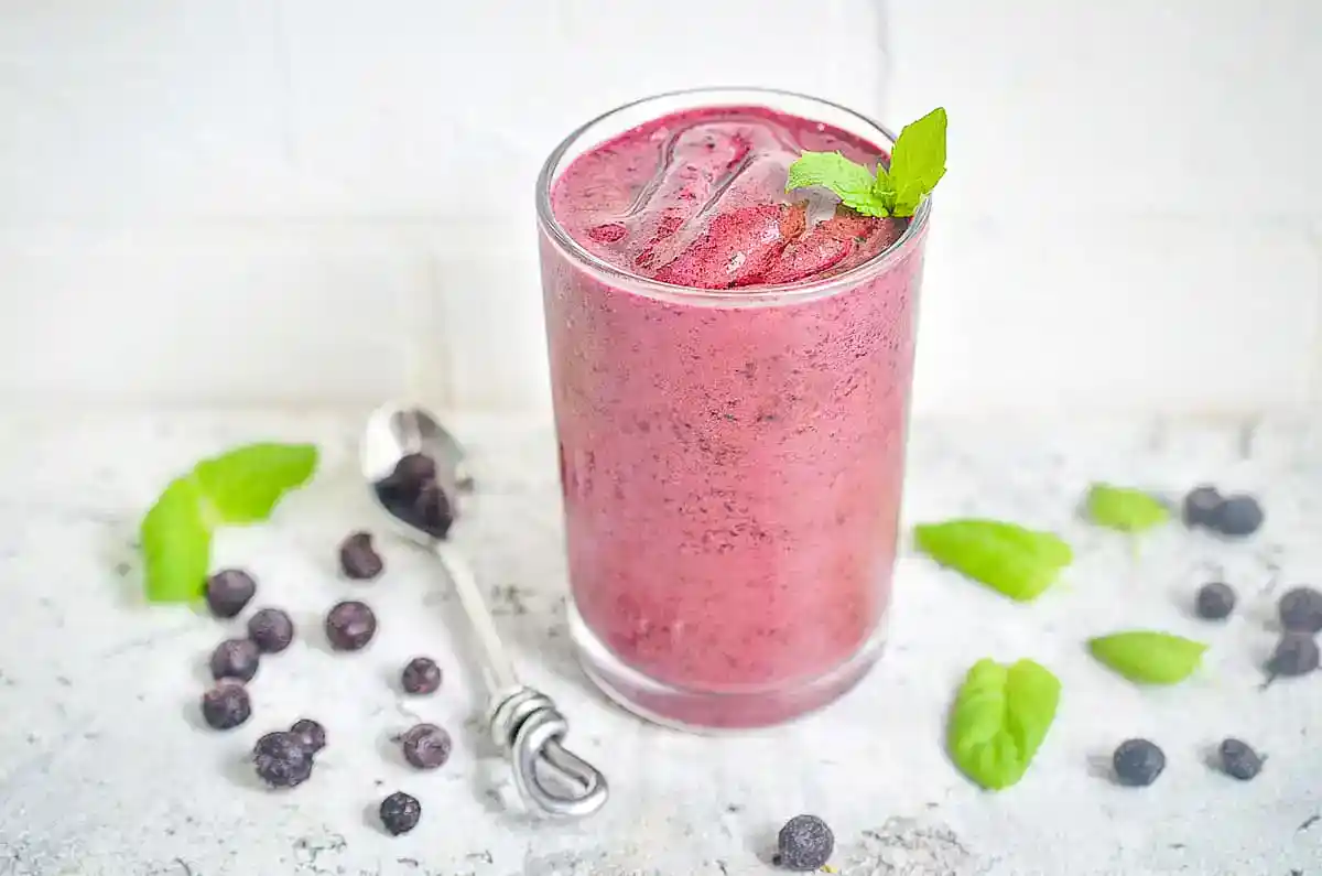 A glass of purple berry smoothie with ice and a mint garnish, surrounded by scattered berries, mint leaves, and a spoon on a light surface.