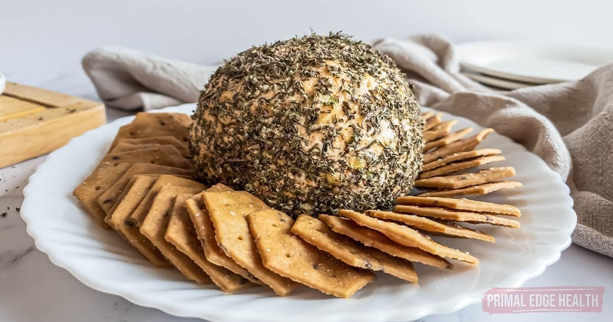 A round cheese ball coated in herbs is served on a white plate surrounded by crackers, with a napkin and stack of plates in the background.