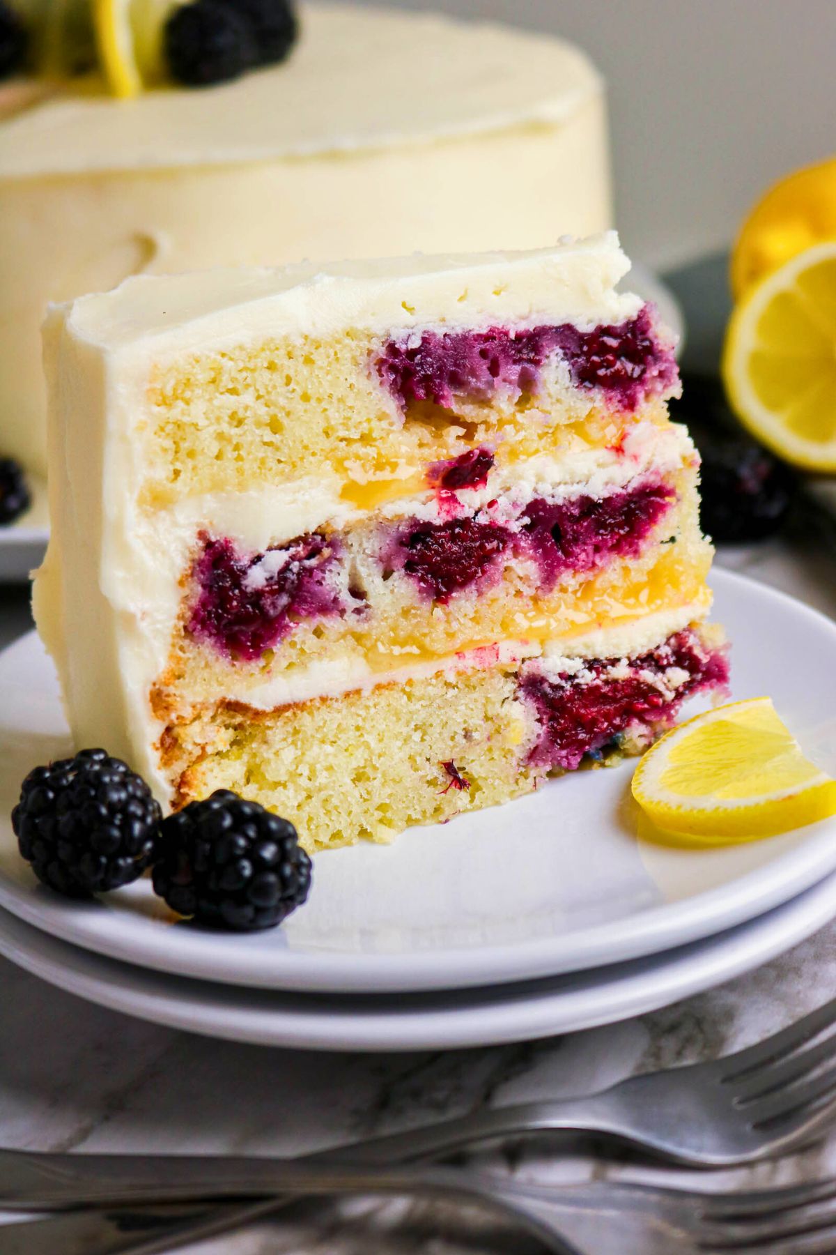 A slice of layered lemon cake with blackberry filling and white frosting on a white plate, garnished with blackberries and a lemon wedge.
