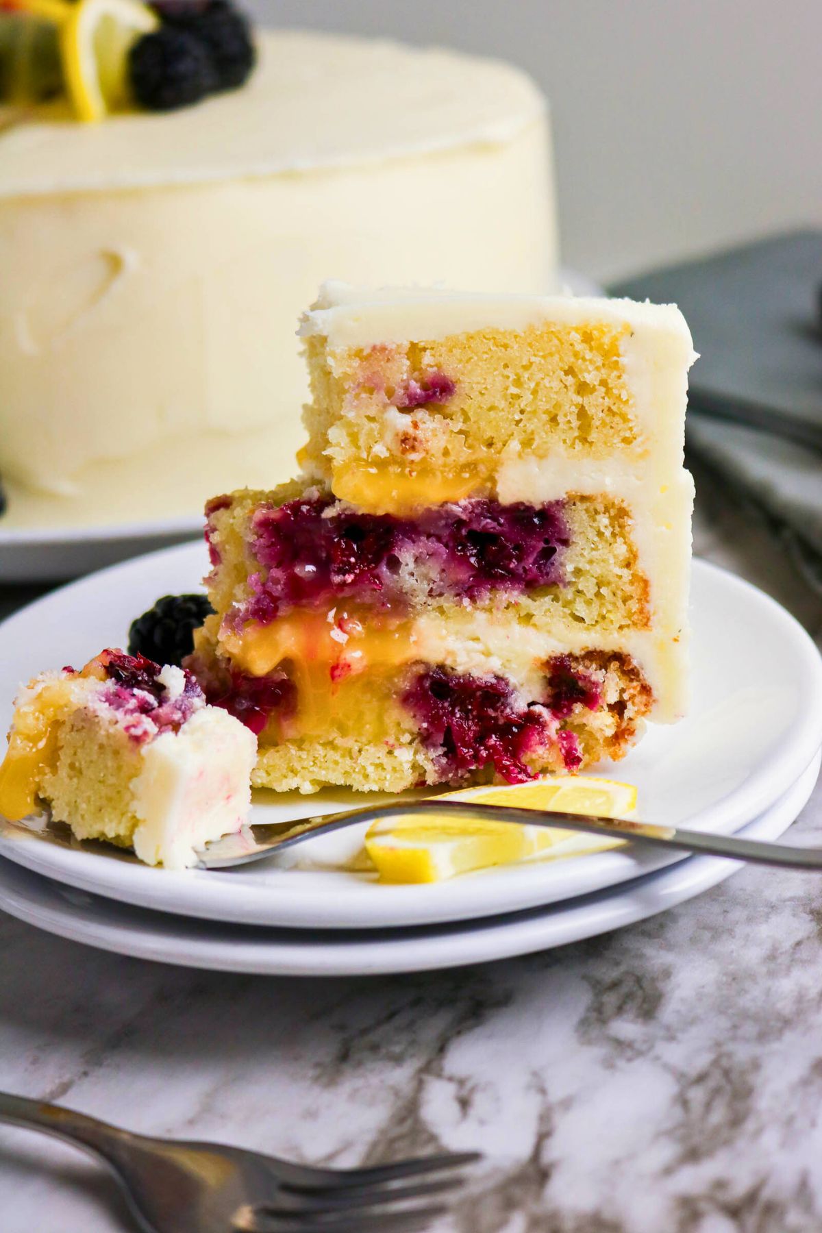A slice of layered cake with white frosting, blackberry and lemon filling, on a white plate with a fork; a whole cake is in the background.