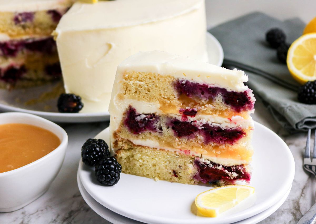A slice of layered cake with berries and cream, garnished with blackberries and a lemon wedge, sits on a white plate beside a bowl of sauce and a whole cake in the background.