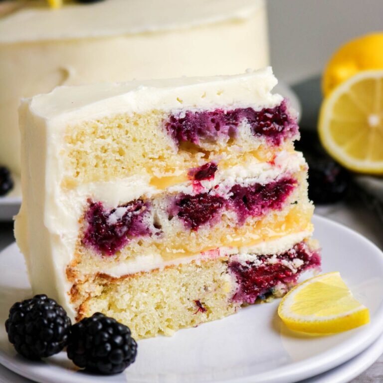 A slice of lemon layer cake with blackberry filling and white frosting on a plate, garnished with fresh blackberries and a lemon slice.