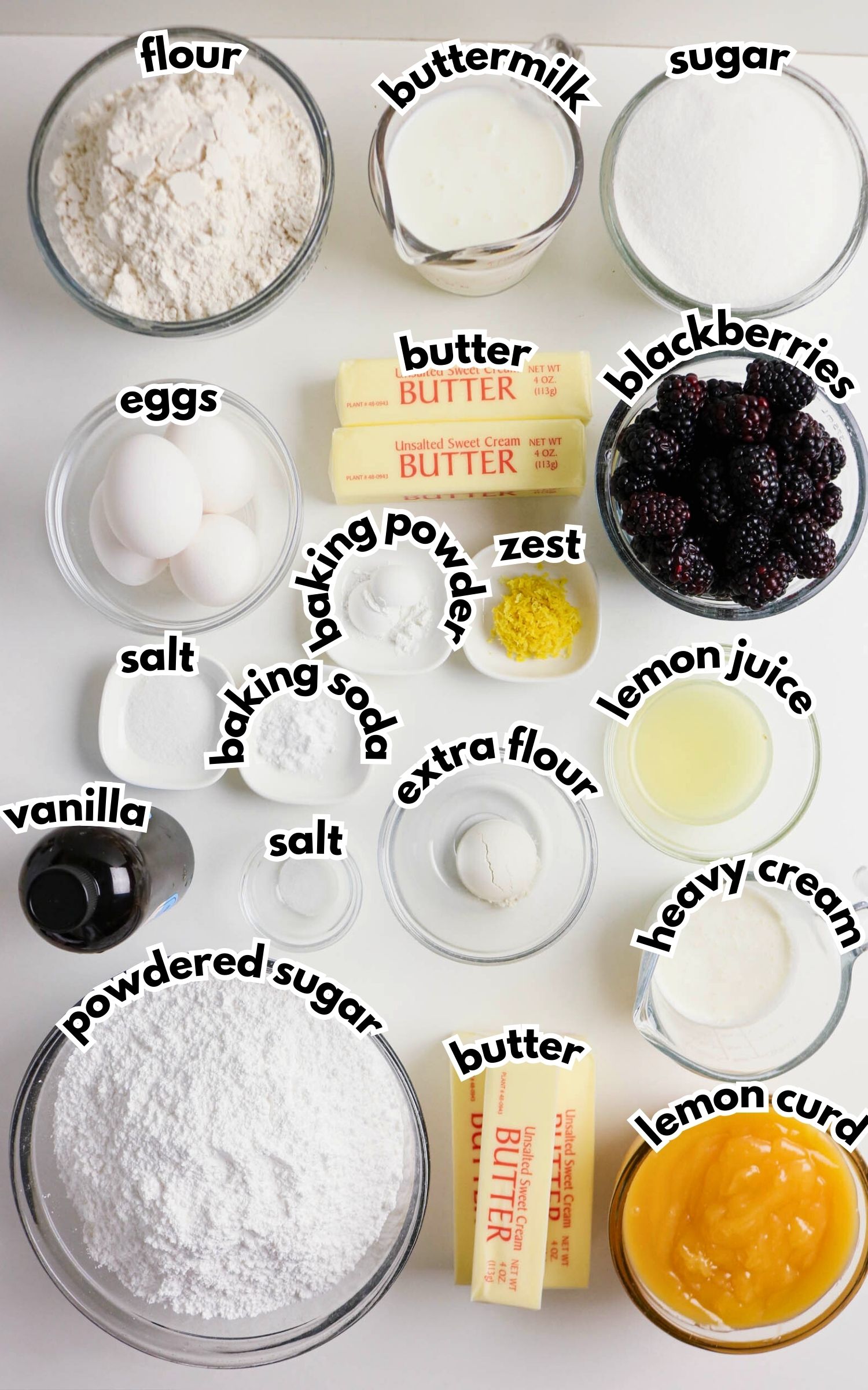 Various baking ingredients in bowls and on plates, including flour, buttermilk, sugar, eggs, butter, blackberries, lemon zest, lemon juice, vanilla, and powdered sugar, labeled with text.