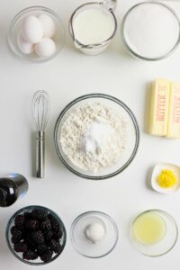 Baking ingredients arranged on a white surface, including eggs, milk, sugar, butter, flour, baking powder, lemon zest, lemon juice, vanilla extract, and blackberries with a whisk.