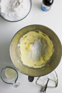 A mixing bowl with butter and powdered sugar, surrounded by a measuring cup of milk, a bowl of flour, and a bottle of vanilla extract on a white surface.