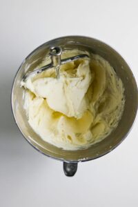 A metal mixing bowl with a paddle attachment contains whipped, pale yellow buttercream on a white surface.