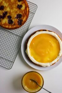 Two cake layers, one with berries cooling on a rack and another on a plate topped with white frosting and yellow curd, with a small bowl of yellow curd and a spoon beside them.