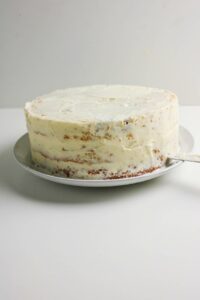A round, partially frosted layer cake sits on a white plate against a plain white background.
