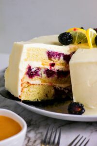 A sliced layered vanilla cake with white frosting, filled with berries and topped with blackberries and lemon slices, sits on a plate next to a bowl of sauce.
