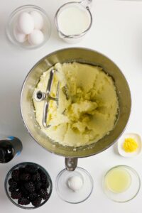 Overhead view of baking ingredients: eggs, milk, butter and sugar creamed in a mixing bowl, lemon zest, lemon juice, blackberries, and vanilla extract on a white surface.