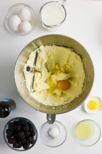 A mixing bowl with creamed butter and sugar and an egg, surrounded by eggs, milk, vanilla, blackberries, lemon juice, zest, and baking powder on a white surface.