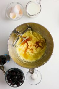 A mixing bowl with creamed ingredients, vanilla extract, and a beater sits on a table alongside eggshells, milk, blackberries, a bottle, and a bowl with one cracked egg.