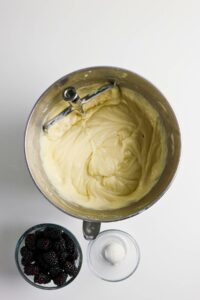A mixing bowl with cake batter and a beater, next to a bowl of blackberries and a small bowl of sugar on a white surface.