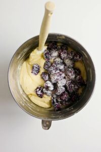 A mixing bowl with cake batter and sugared blackberries, with a wooden spoon resting inside.