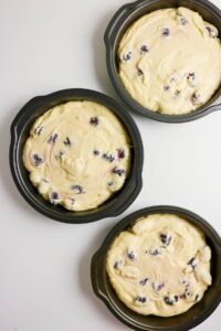 Three round cake pans filled with unbaked batter mixed with blackberries are arranged on a white surface.