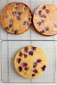 Three round cake layers with purple spots are cooling on wire racks, each layer covered with a thin sheet of parchment paper.