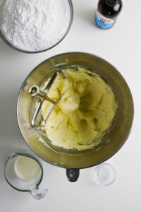 A mixing bowl with creamed butter, a paddle attachment, and surrounding ingredients: powdered sugar, vanilla extract, milk, and a small bowl of salt on a white surface.