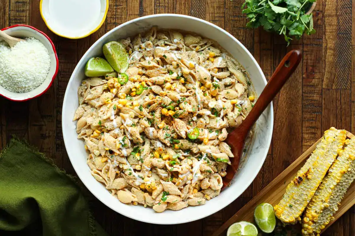 A large bowl of pasta shells mixed with corn, herbs, and sauce, garnished with lime wedges, next to grilled corn, lime halves, grated cheese, and fresh herbs on a wooden table.