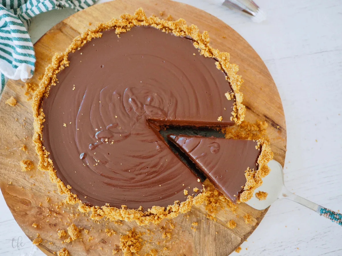 A chocolate tart with a graham cracker crust, one slice being removed with a pie server, on a wooden cutting board.