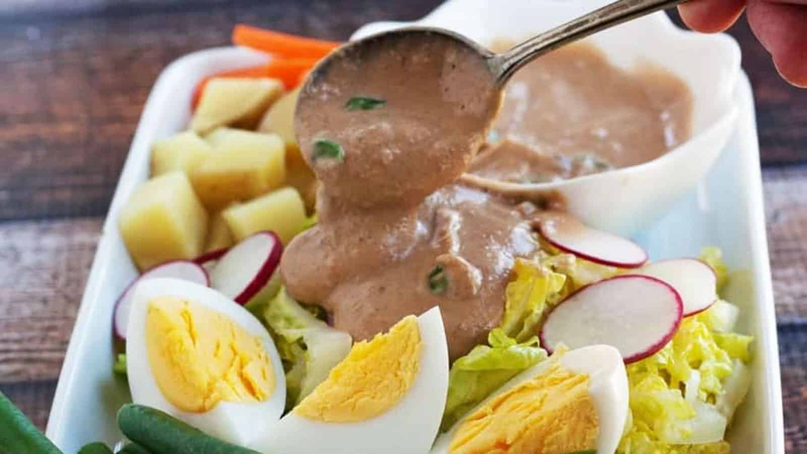 A hand pours brown peanut sauce over a salad with lettuce, sliced boiled eggs, radishes, potatoes, carrots, and green beans on a white plate.