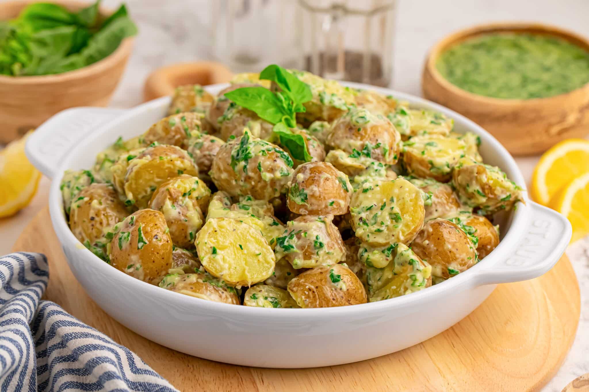 A white dish filled with creamy potato salad garnished with fresh herbs, placed on a wooden board with lemons, sauce, and a striped napkin nearby.