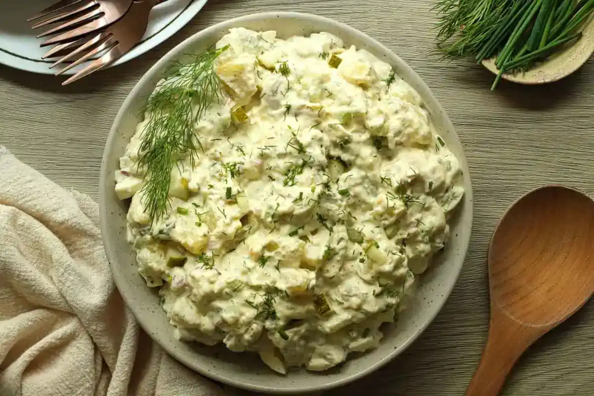 A bowl of creamy potato salad garnished with fresh dill sits on a wooden table beside a wooden spoon and a plate with a fork and knife.