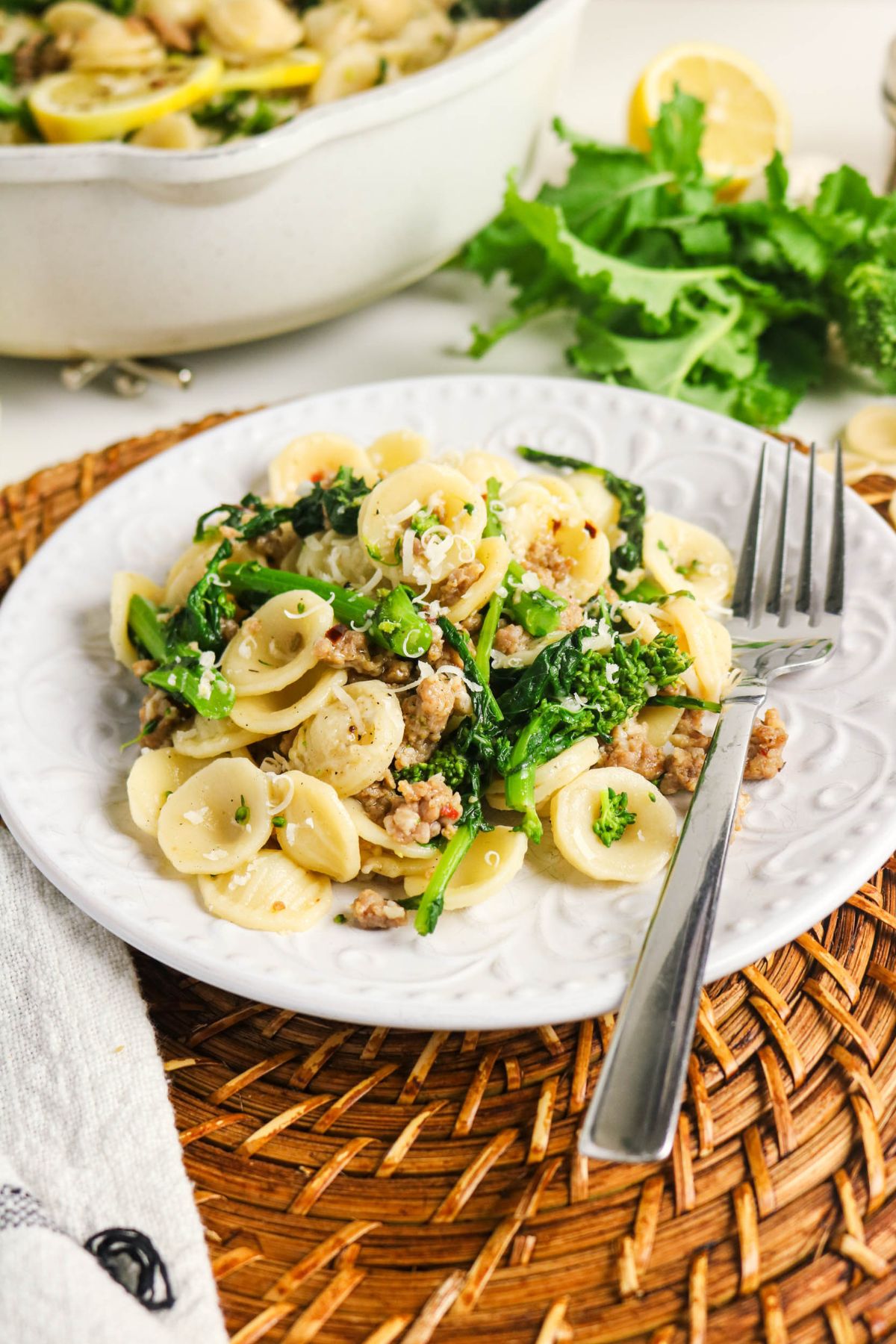 A plate of orecchiette pasta with sausage, leafy greens, and grated cheese, served on a white plate with a fork on a woven placemat.