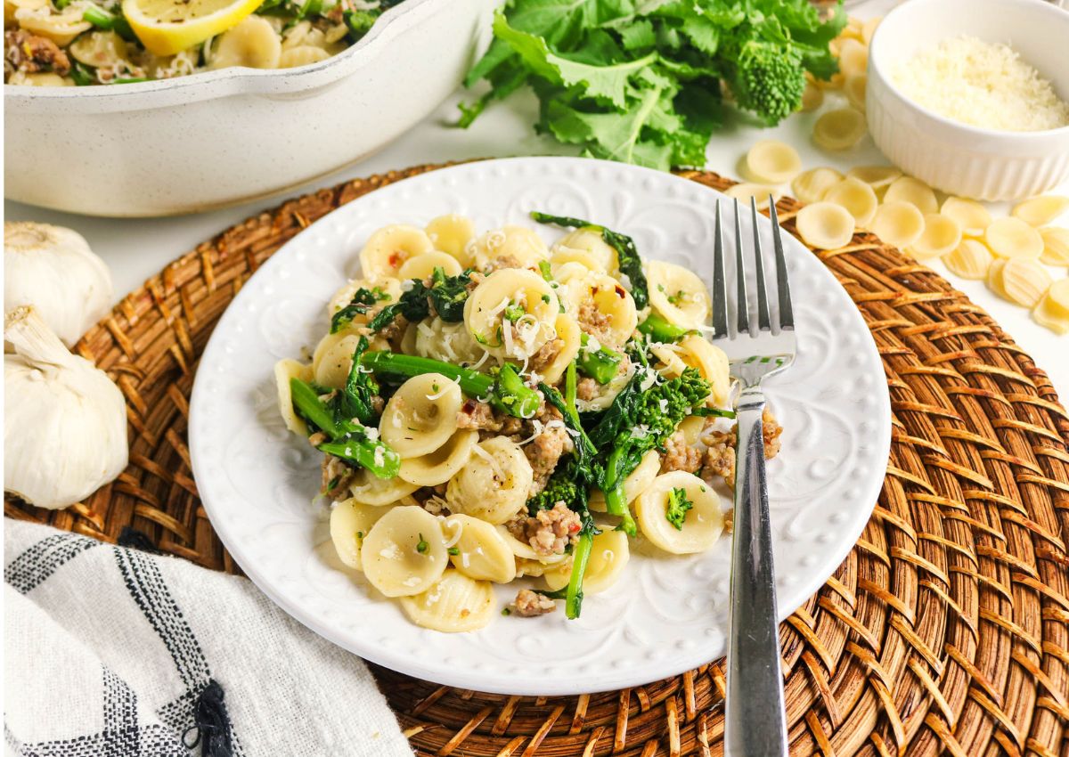 A plate of orecchiette pasta with sausage and broccoli rabe, garnished with herbs, sits on a white plate with a fork, next to a dish of grated cheese and fresh ingredients.