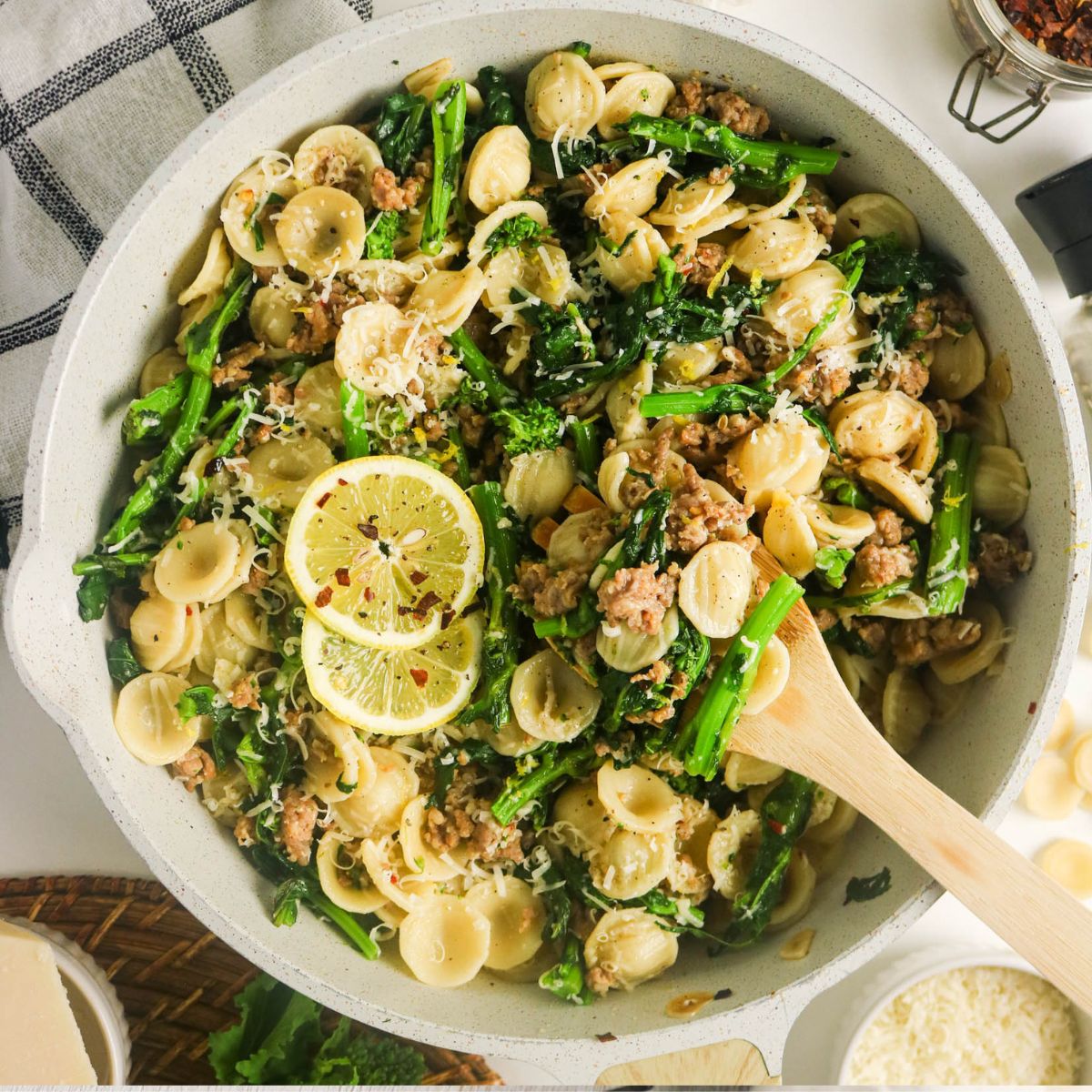 Orecchiette pasta with sausage, broccoli rabe, lemon slices, and grated cheese in a white skillet, stirred with a wooden spoon.