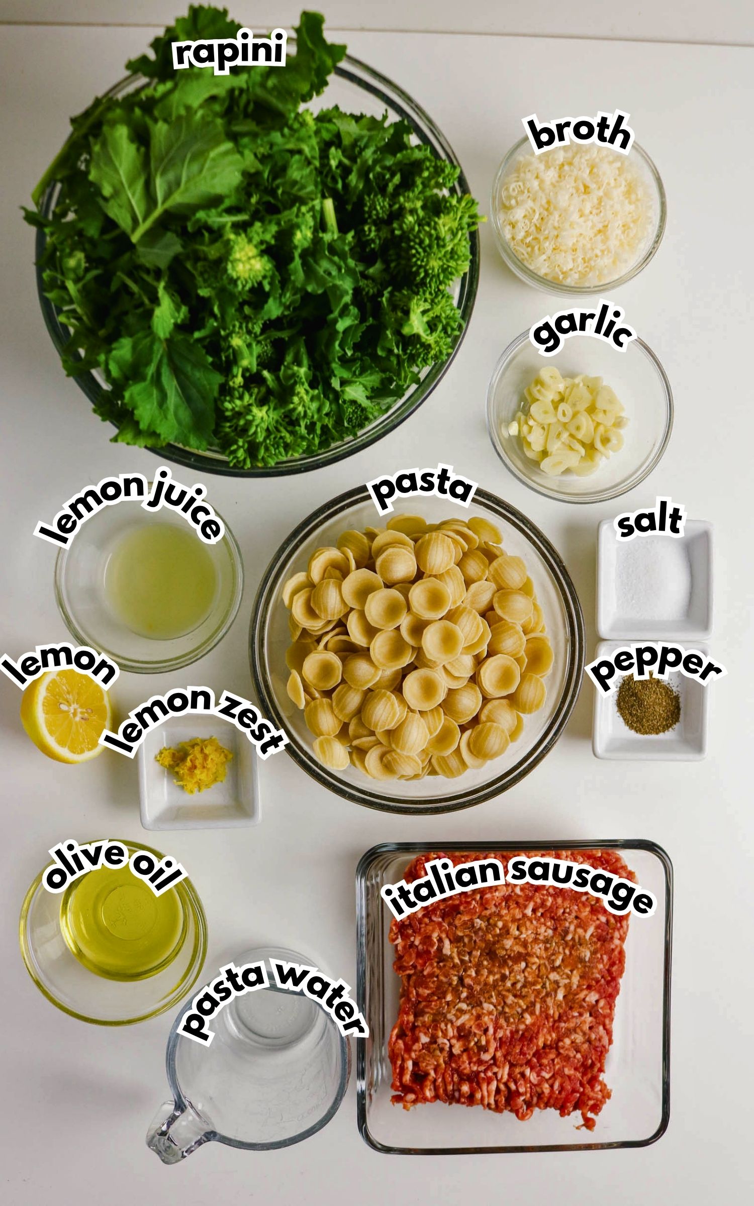 A flat lay of ingredients for pasta with rapini and sausage, including rapini, orecchiette pasta, Italian sausage, garlic, broth, lemon, olive oil, salt, pepper, pasta water, and grated cheese.