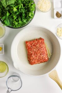 Ground meat is cooking in a white skillet with oil, surrounded by bowls of chopped greens, grated cheese, sliced garlic, seasonings, and liquids on a white countertop.