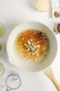 A white pan with sliced garlic sautéing in oil, showing browned bits; surrounding are bowls of grated cheese, spices, and liquids on a white surface.