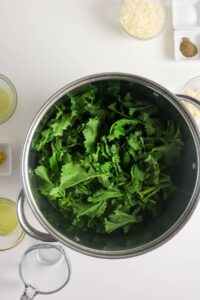 A large pot filled with fresh kale leaves on a white surface, surrounded by small bowls containing various ingredients such as grated cheese, spices, and liquids.