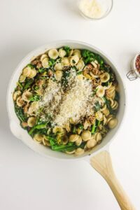 A skillet containing orecchiette pasta with broccoli, sausage, and shredded cheese on top, placed on a white surface.