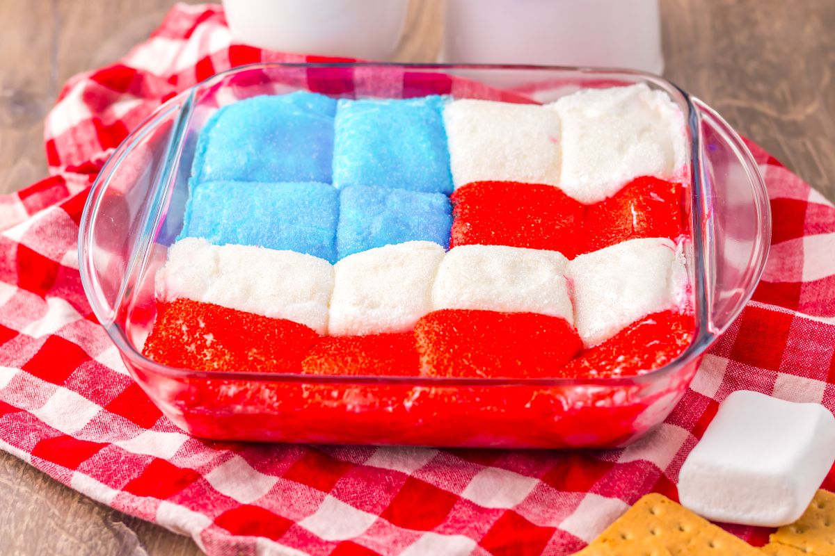 A glass dish contains marshmallows arranged and colored to resemble the American flag, set on a red checkered cloth with graham crackers nearby.