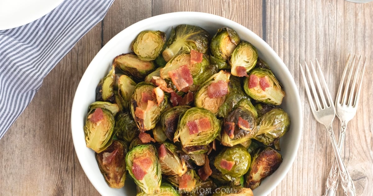 A white bowl filled with roasted Brussels sprouts topped with pieces of bacon, placed on a wooden table with two forks beside it.