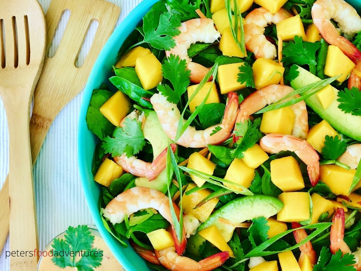 A bowl of salad with shrimp, mango cubes, avocado slices, fresh greens, and cilantro, next to wooden utensils.