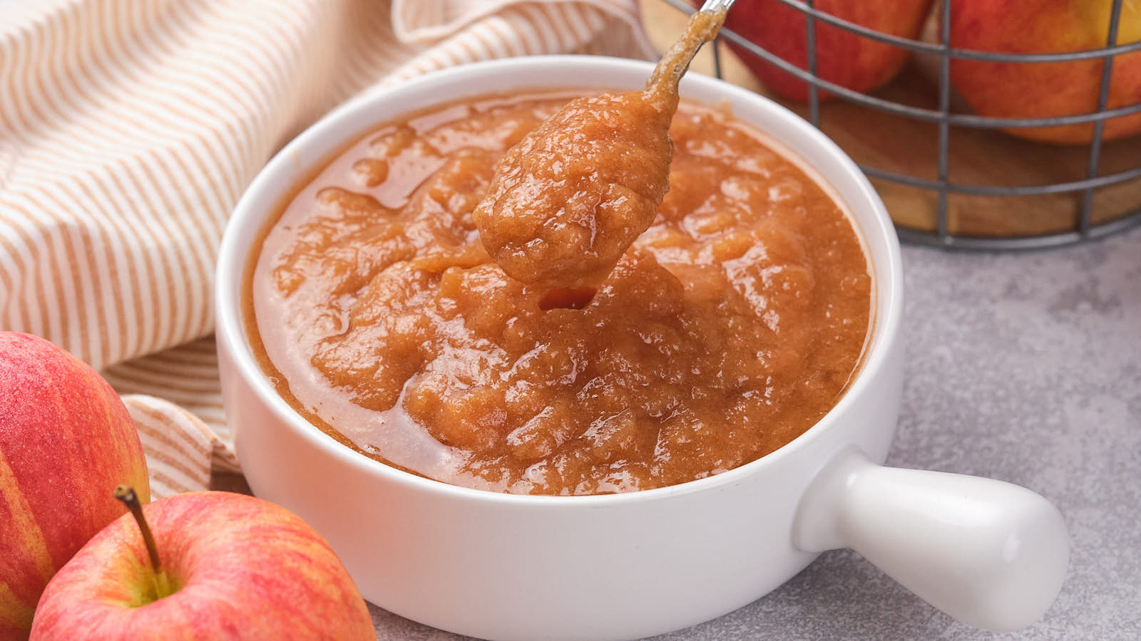A white bowl filled with applesauce sits on a gray surface, with a spoon scooping some out. Fresh apples and a wire basket of apples are nearby.