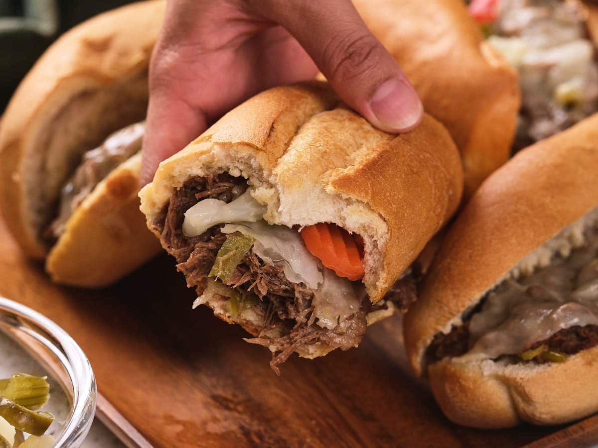A hand holding a sandwich filled with shredded beef, melted cheese, and vegetables, with more sandwiches on a wooden surface in the background.