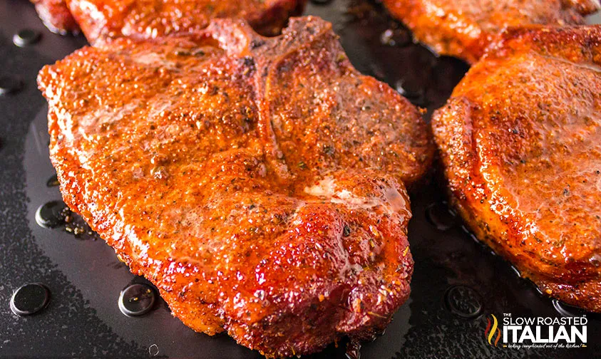 Close-up of seasoned, roasted pork chops on a dark surface, with visible juices and a logo in the bottom right corner reading The Slow Roasted Italian.
