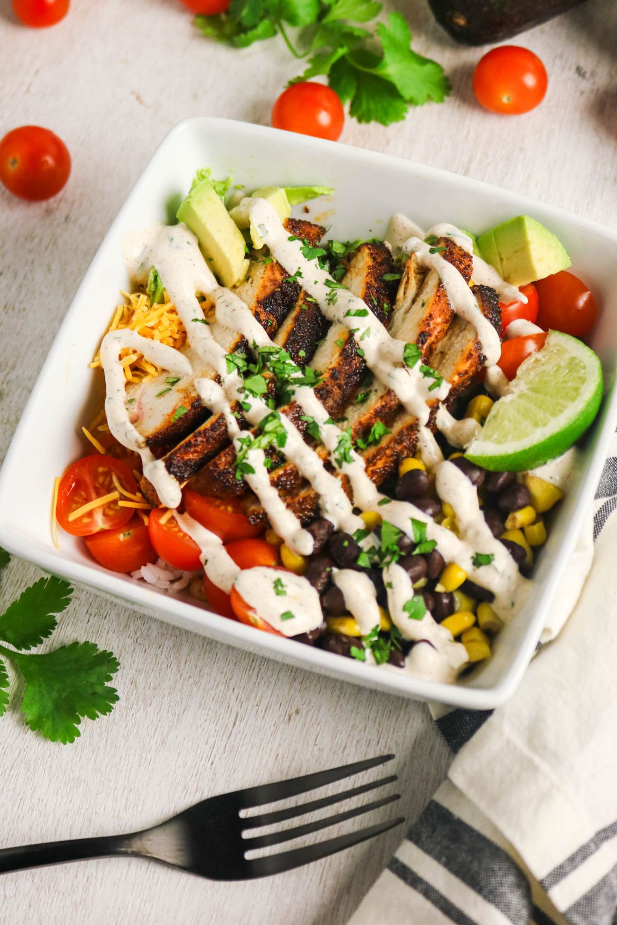 A square bowl with sliced grilled chicken, black beans, corn, shredded cheese, cherry tomatoes, avocado, and lime, drizzled with a creamy sauce and garnished with chopped herbs.