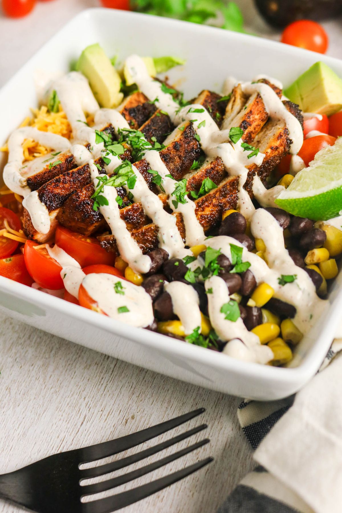 A bowl filled with grilled chicken, black beans, corn, cherry tomatoes, avocado, shredded cheese, and lime, topped with a creamy white sauce and garnished with chopped herbs.