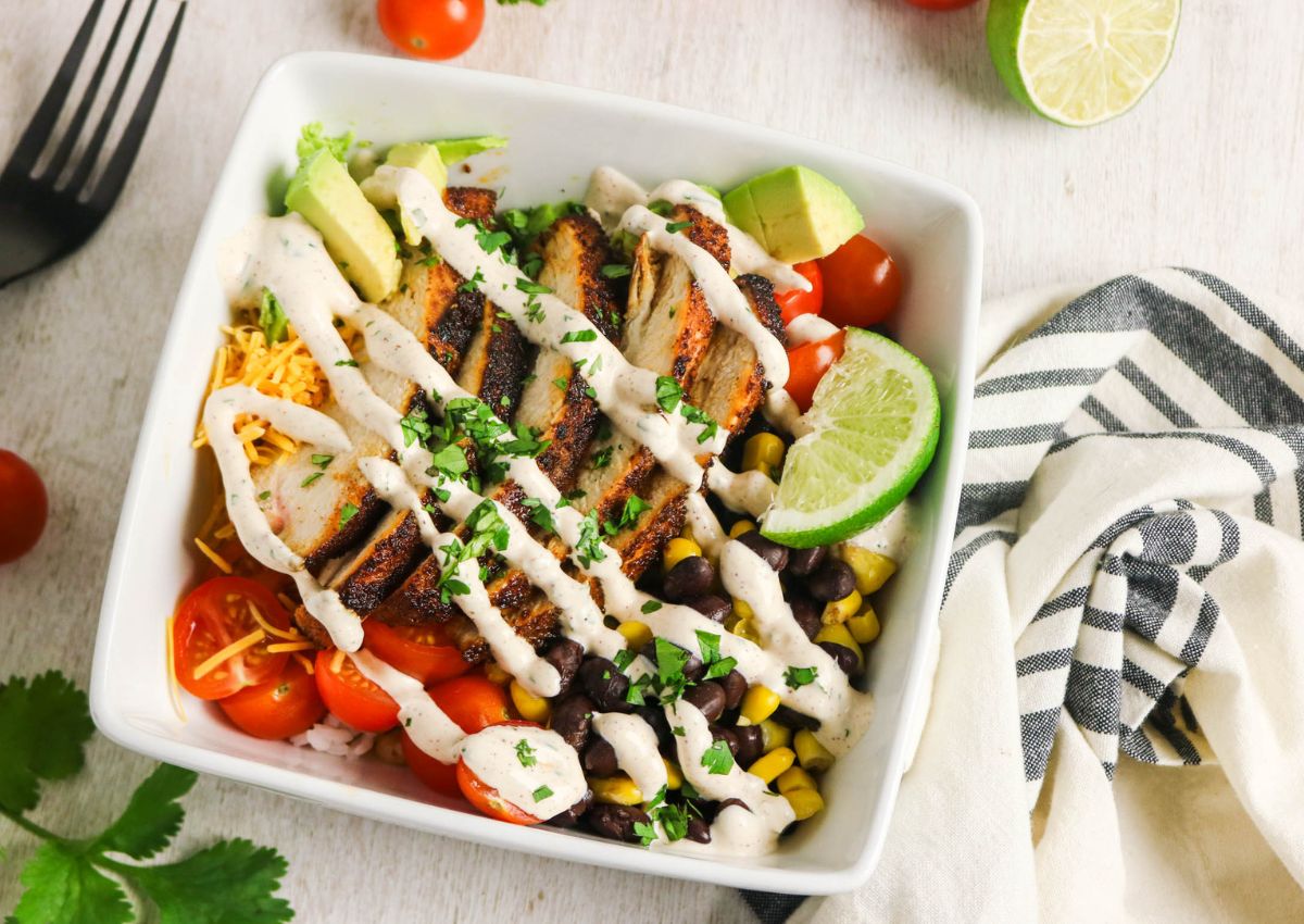 A bowl containing grilled chicken, cherry tomatoes, black beans, corn, avocado, shredded cheese, and a lime wedge, topped with creamy dressing and chopped herbs.