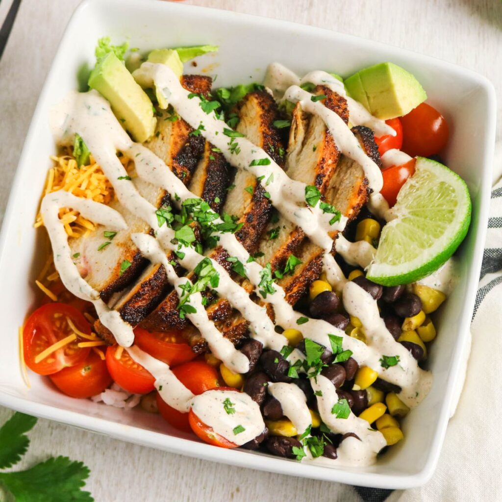A bowl with grilled chicken slices, cherry tomatoes, black beans, corn, shredded cheese, avocado chunks, a lime wedge, and creamy dressing, garnished with chopped herbs.
