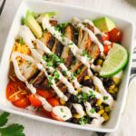 A bowl with grilled chicken slices, cherry tomatoes, black beans, corn, shredded cheese, avocado chunks, a lime wedge, and creamy dressing, garnished with chopped herbs.