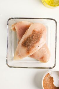 Two raw chicken breasts in a glass dish with a pile of seasoning on top; a bowl of seasoning and a container of oil are nearby.