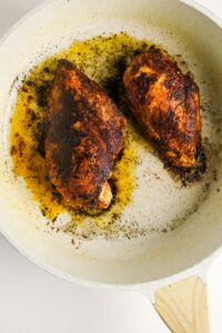 Two seasoned and cooked chicken breasts in a white skillet with oil and browned spices visible around the meat.