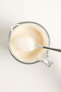 A glass measuring cup filled with creamy white sauce, with a spoon resting inside and some sauce on the spoon.