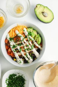A bowl of salad with chicken, black beans, corn, cherry tomatoes, shredded cheese, avocado slices, and dressing, surrounded by avocado, herbs, and small bowls of ingredients on a white surface.