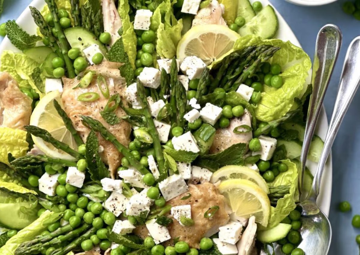 A salad with lettuce, green peas, asparagus, cucumber slices, lemon slices, feta cheese cubes, chicken pieces, and mint leaves, served with two spoons.